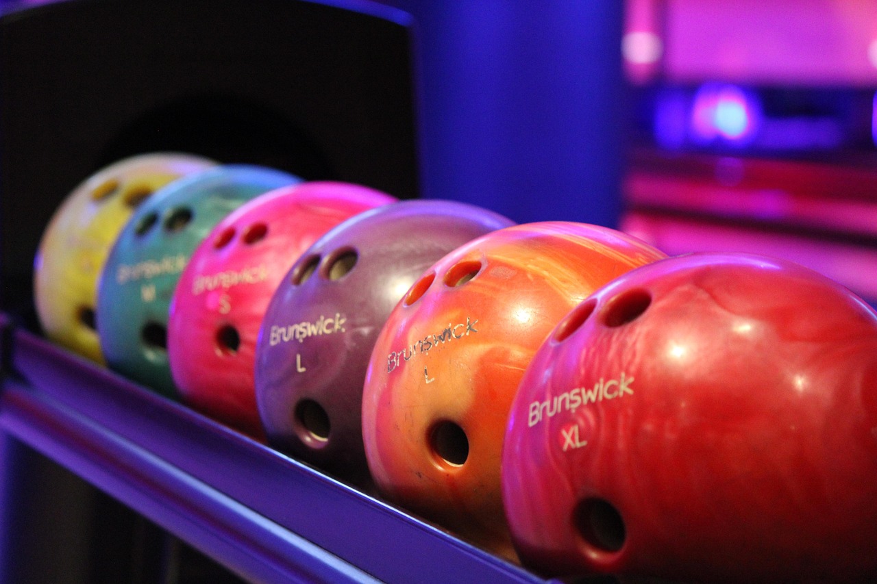 I en mörk lokal ligger sex bowlingklot i olika färger. I bakgrunden lysen en lila lampa.