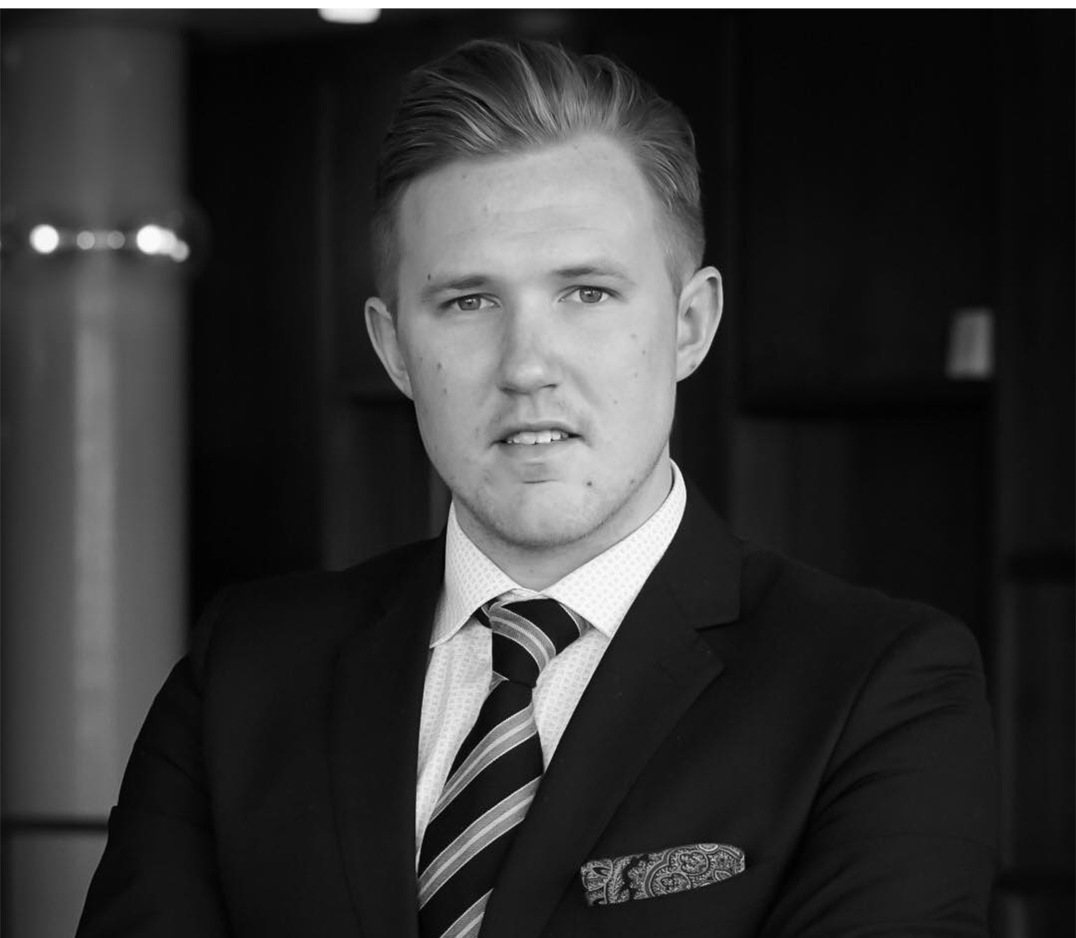 I en svartvit bild står en man i kostym med händerna i kors och tittar in i bild.
