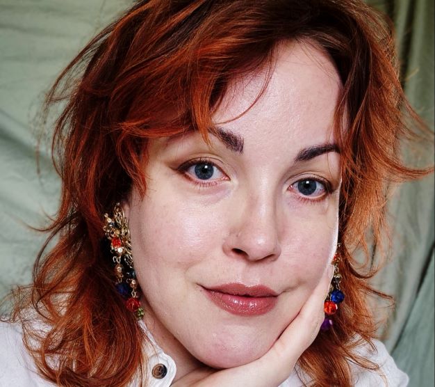 En rödhårig kvinna med rödmålade läppar, bär en vit blus. Hon vilar huvudet mot ena handen och ler mot betraktaren. 
