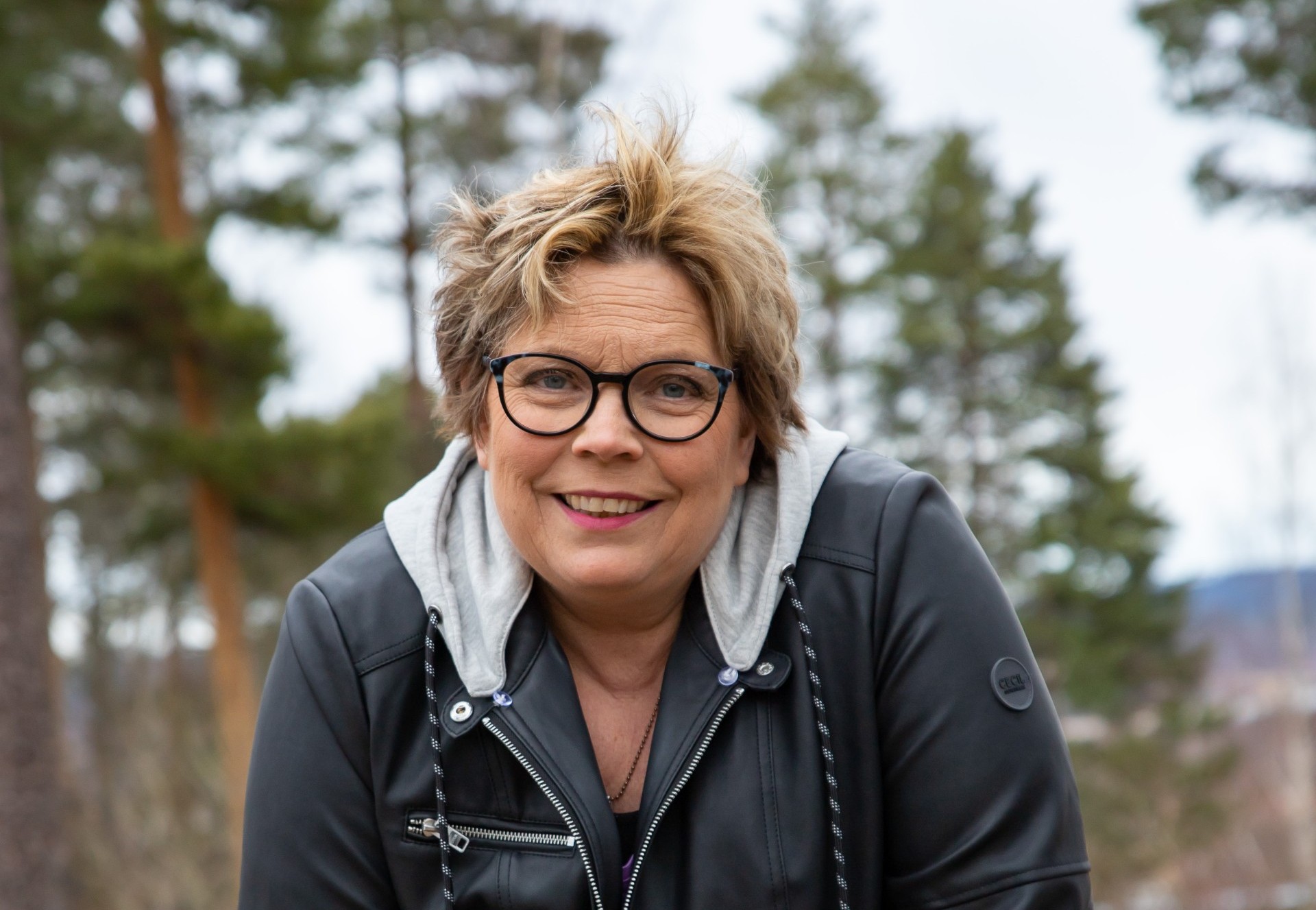 En leende kvinna med vindrufs i håret lutar sig framåt. Hon har skinnjacka och hoodie på sig. 