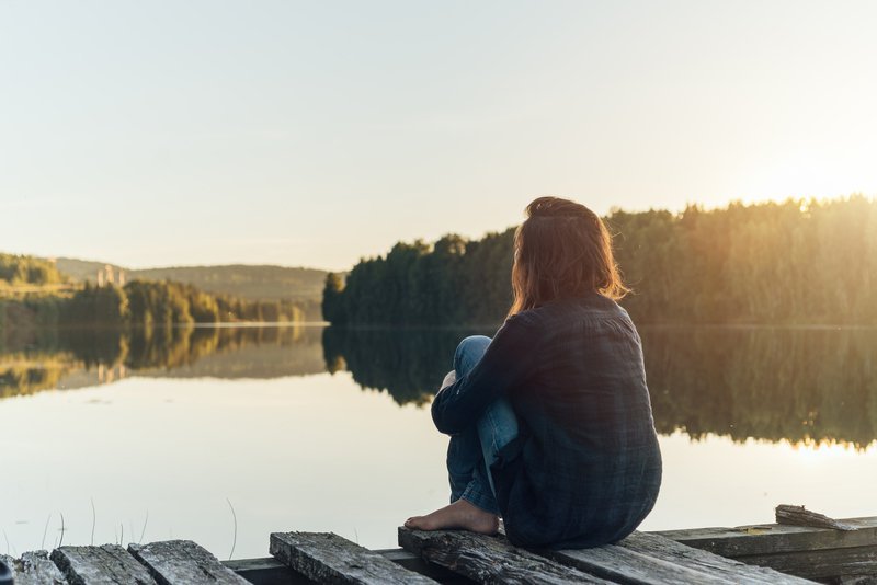 En kvinna sitter ihopkrupen på en brygga vid en sjö med skogsklädda öar.
