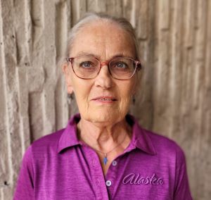 Kerstin, en äldre kvinna med grått uppsatt hår. Hon har en lila topp och matchande glasögon. Runt halsen bär hon ett blått kors.