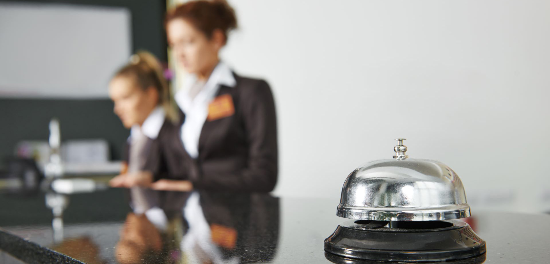 En ringklocka på en receptionsdisk. I bakgrunden jobbar två kvinnor.