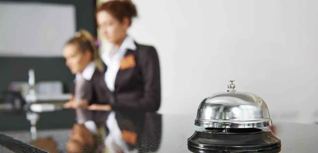 En ringklocka på en receptionsdisk. I bakgrunden jobbar två kvinnor.