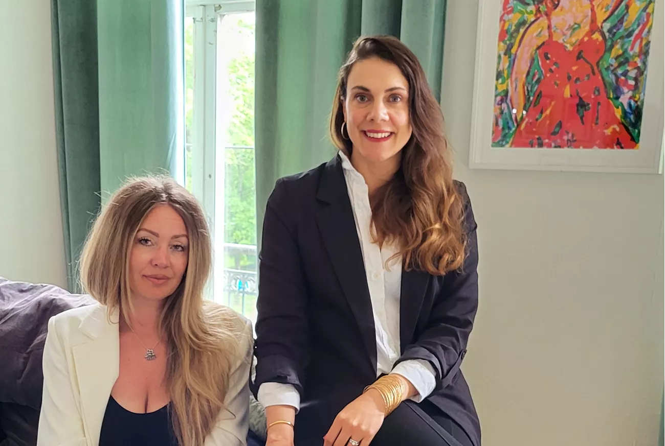 En blond kvinna i vit kostym sitter i en soffa och på armstödet sitter en mörkhårig kvinna i svart kostym . Text: För livet - tillsammans med Johanna och Maria.